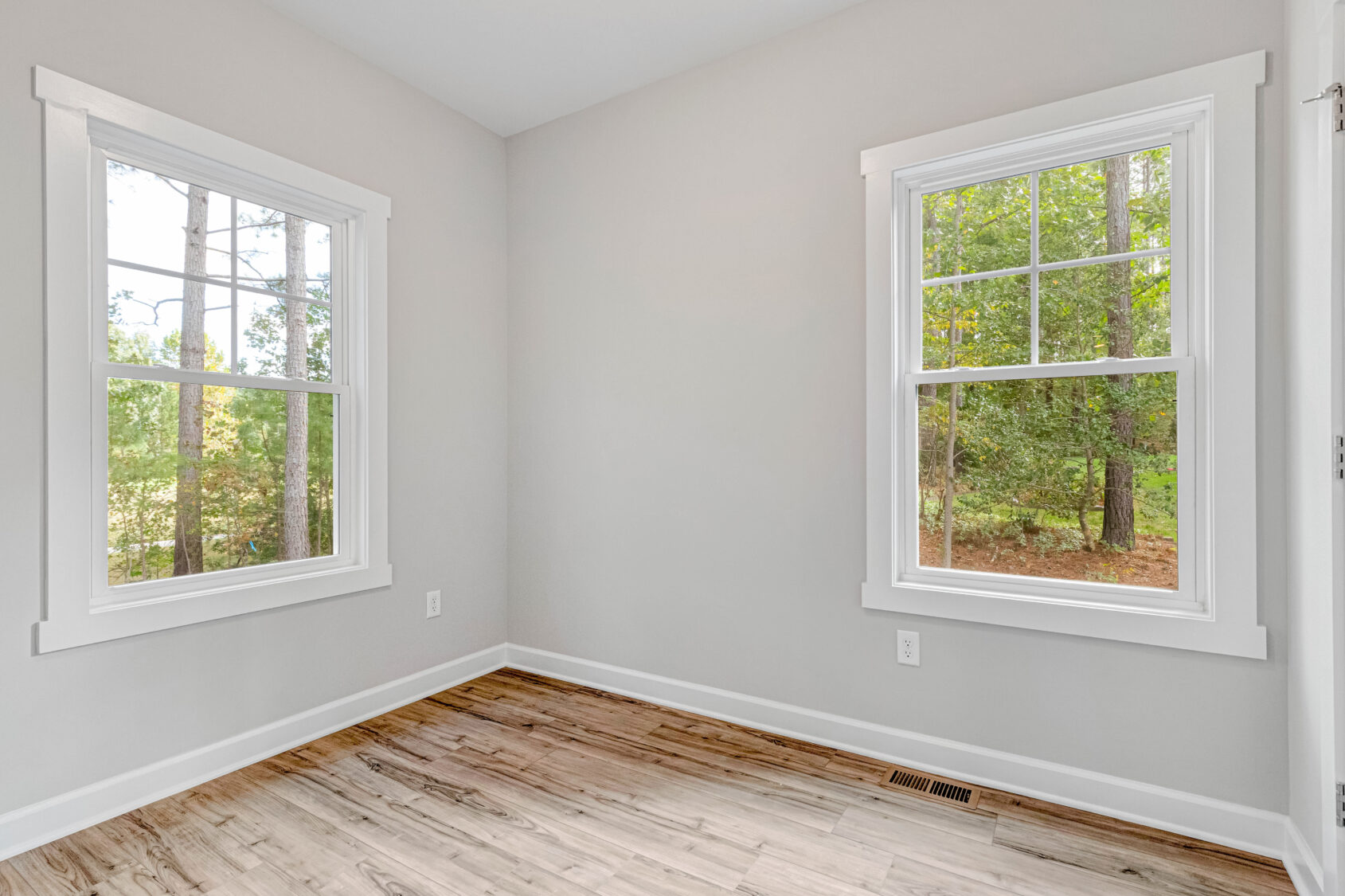 Empty room with light gray walls, two large windows, and a wood floor, overlooking trees outside.