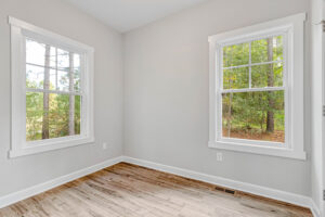 Empty room with light gray walls, two large windows, and a wood floor, overlooking trees outside.