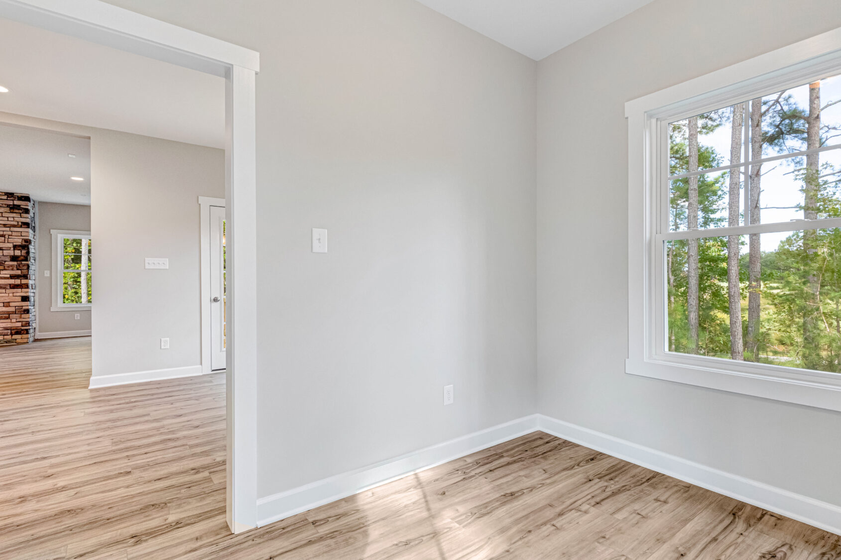 Bright empty room with wood floors, light walls, and a large window overlooking green trees.