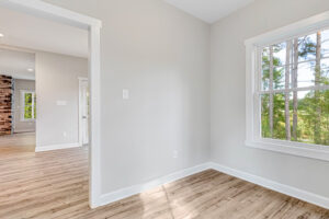 Bright empty room with wood floors, light walls, and a large window overlooking green trees.