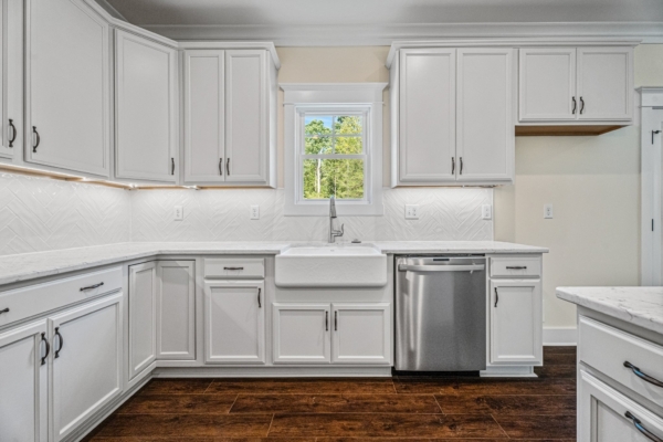A modern kitchen in a new home with white cabinets, a farmhouse sink, stainless steel dishwasher, and dark wood flooring.