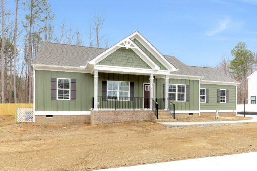 Single-story green house with a ranch plan, front porch, white railing, and a dirt yard surrounded by sparse trees.