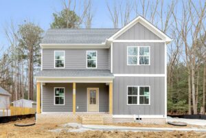 Two-story gray house with a screen porch under construction, surrounded by trees and a bare dirt front yard.