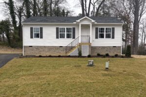 Single-story house with white siding and brick foundation on Lot 15, Poplar Village. Grassy lawn and surrounding trees.