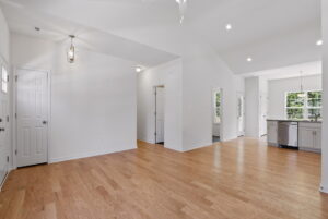 Bright, empty room with light wood floors, white walls, and open entry to kitchen and dining area.