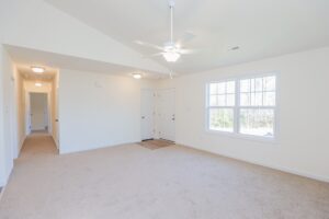Bright, empty room with beige carpet, ceiling fan, large window, and double doors near the entrance.