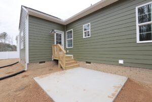 Small wooden steps lead to the back door of this ranch plan with a green house, concrete patio, and dirt yard.