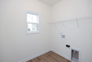 Empty laundry room in new homes in Richmond VA with white walls, wire shelf, window, and washer/dryer hookups.