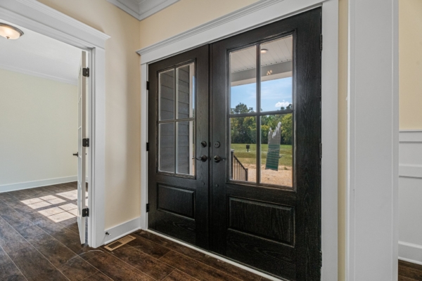 Black double doors with glass panels open to a grassy view in this sunlit new home. Wooden floors and white walls inside.