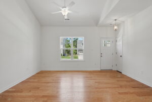 Bright empty room with light wood floors, white walls, ceiling fan, window, and front door with glass panes.