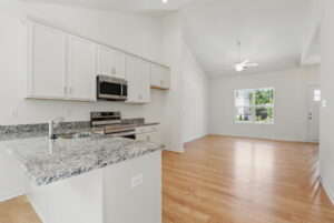 Modern kitchen with white cabinets and granite countertops opens to a bright, empty living room with hardwood floors.