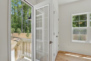 Glass door opens to a wooden deck in Poplar Village, with fenced backyard and green trees under a blue sky.