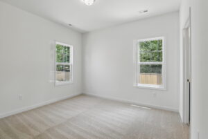 Bright, empty room with beige carpet, white walls, and two windows facing a wooden fence and trees.
