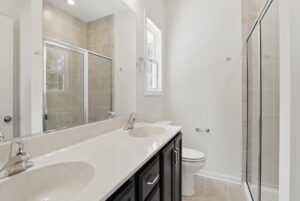 Modern bathroom with double sinks, a glass shower, toilet, large mirror, and beige tiled walls and floor.