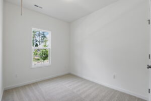 Empty room with white walls, beige carpet, and a window showing trees and blue sky outside.