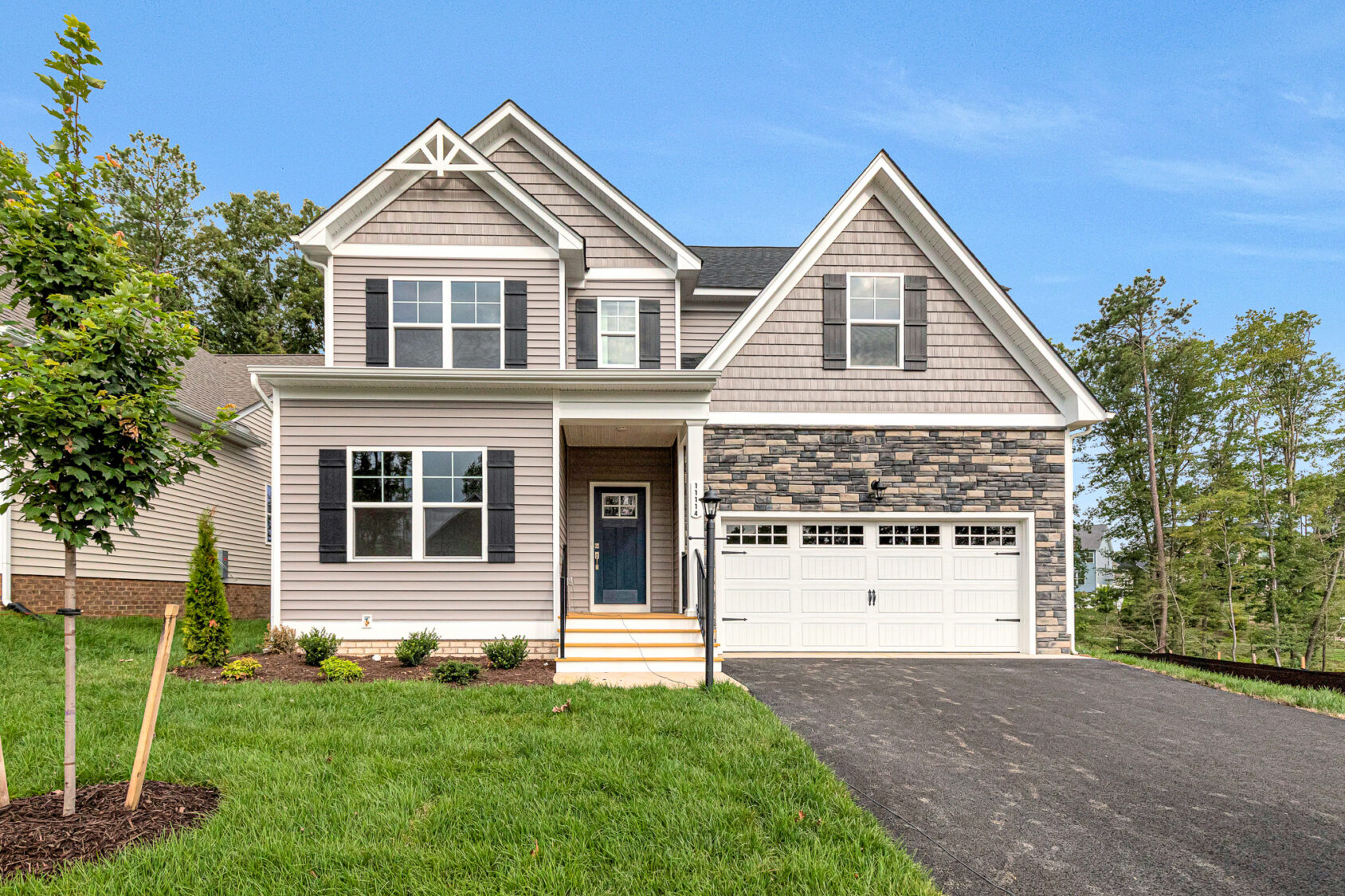 Two-story Somerset Hill home with gray siding, stone accents, two-car garage, and lush grass surrounded by trees.