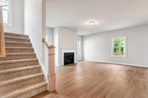 Bright, empty Somerset Hill living room with wood floors, fireplace, staircase, and windows showing greenery outside.