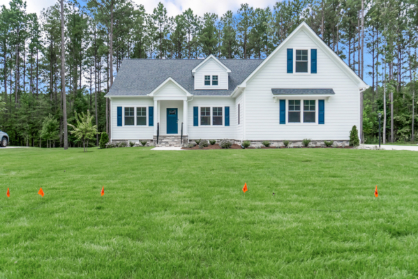A white house with blue shutters sits in front of a forest, on a grassy lawn adorned with small orange flags—a perfect new home.