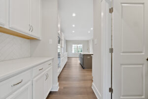 Modern kitchen with white cabinets, herringbone backsplash, and wood flooring in a new home, leading to a room with large windows.