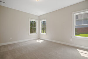 Empty room with beige walls and carpet, two windows letting in natural light, and a view of grass and trees outside.