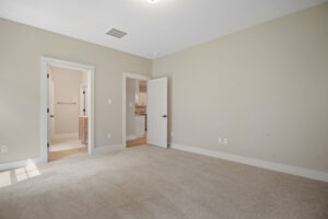 Empty beige carpeted room with a door leading to a bathroom and an open door to a kitchen.