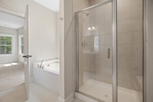 Modern bathroom with glass shower, white bathtub, and open door leading to a bright carpeted room.