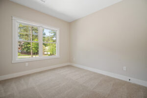 Empty room with beige walls, carpeted floor, and a large window showing trees outside.