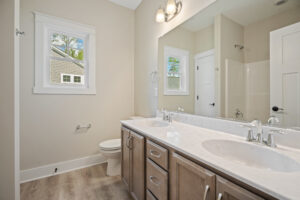 Modern bathroom with double sinks, large mirror, toilet, and window. Neutral tones and wooden cabinets.