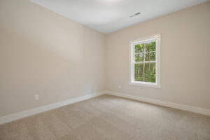 Empty room with beige walls, carpeted floor, and a window showing greenery outside.