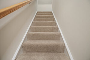 Carpeted staircase with a wooden handrail on the left, leading upwards between light-colored walls.