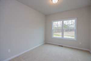 Empty room in new homes in Richmond VA, with beige walls, carpet, ceiling light, and a window showing trees outside.