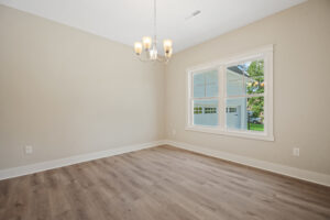 Empty room with beige walls, wooden floor, chandelier, and a window showing trees and a garage outside.