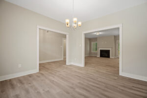 Spacious empty room with light wood flooring, beige walls, and a chandelier, leading to rooms with a fireplace.