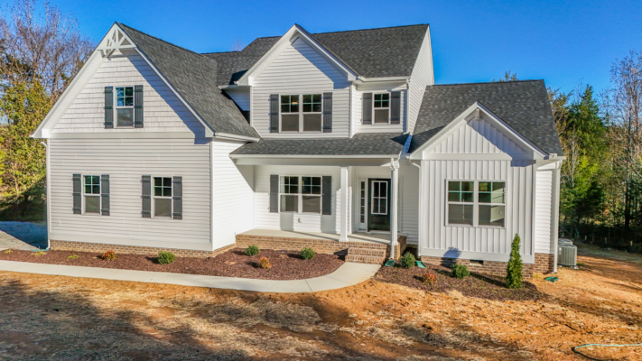 A modern, two-story white house with a dark roof and front porch, looking like a photo gallery amid bare earth and scattered trees.