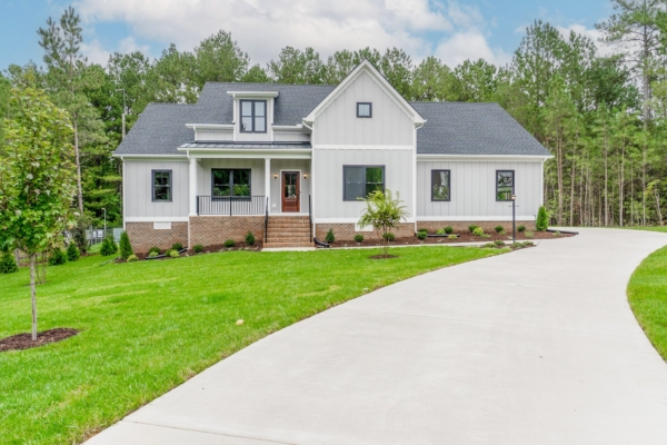 Modern white house with black roof, surrounded by trees, a green lawn, and a curved driveway leading to the entrance.