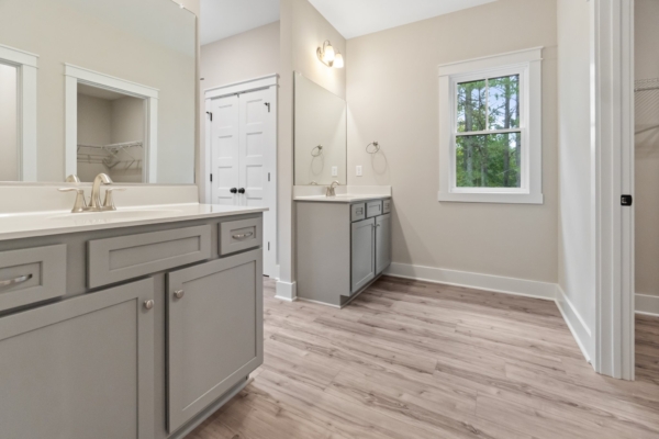 Modern bathroom with dual gray vanities, cream walls, wood floor, and a window with a forest view.