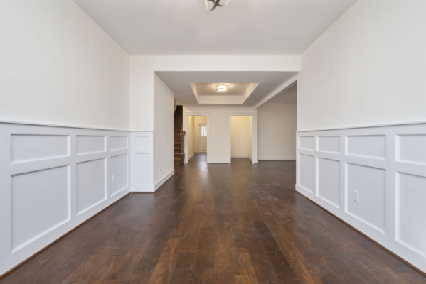Spacious empty room with wooden floors, white wainscoting, recessed ceiling light, and staircase in the background.