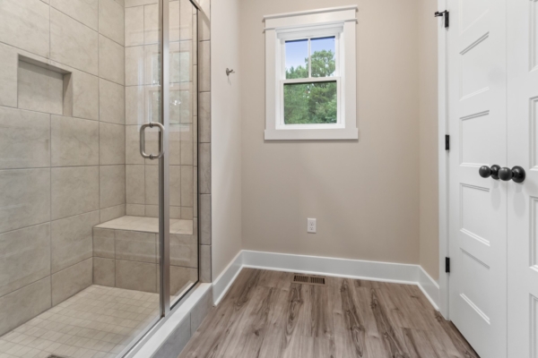 Minimalist bathroom with glass shower, beige walls, a window, and wooden floor.