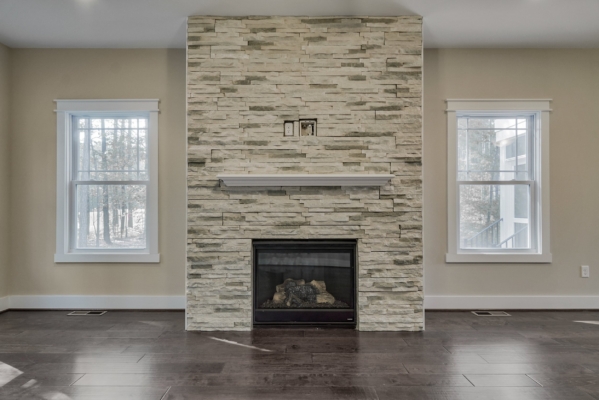 Stone fireplace with a mantel between two windows in a living room with dark wood floors.