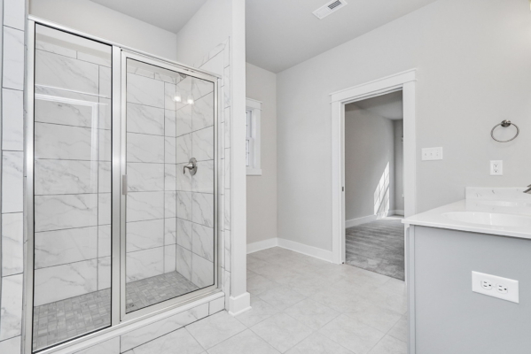 Modern bathroom with glass shower, white tiled walls, and a light gray vanity next to an open doorway.