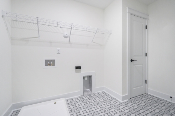 Minimalist laundry room with patterned floor tiles, wire shelf, and utility hookups on a white wall.