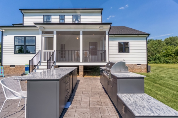 Outdoor kitchen with gray cabinets, marble countertops, and a grill, in front of a white house with a screened porch.