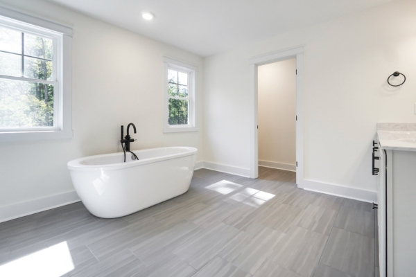 Minimalist bathroom with freestanding tub, gray tile floor, two windows, and an open doorway.