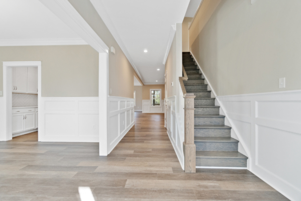 Modern hallway with wooden flooring, beige walls, and a staircase. Doorway leads to a bright kitchen.
