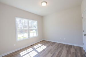 Empty room with light wooden floor, large window, white walls, and a ceiling light. Natural light fills the space.