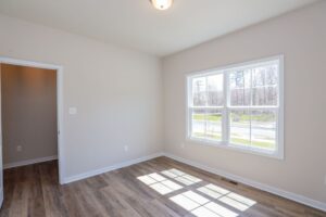 Empty room with a large window, wooden floor, and neutral walls, illuminated by natural light. Doorway visible on the left.
