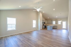 Spacious, empty modern living room with wooden floors, ceiling fan, and view of open kitchen area.