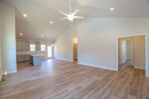 Spacious empty room with wooden flooring, ceiling fan, and view of a kitchen area with an island and overhead lights.