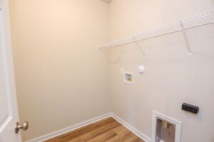 Empty laundry room with beige walls, wood flooring, a wire shelf, and utility hookups.