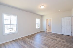 Empty room with light wood flooring, beige walls, ceiling light, two windows, and a door leading to another room.